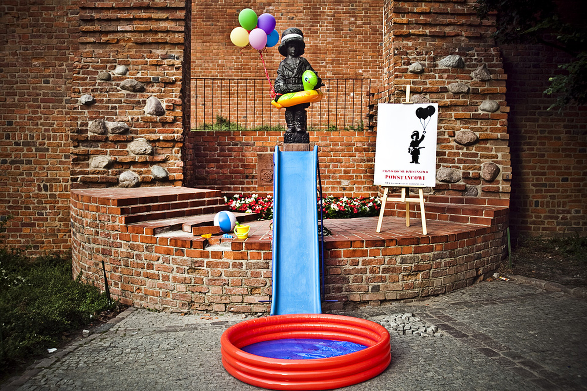  Zdjęcie przedstawia pomnik Małego Powstańca przy warszawskiej starówce. Wokół pomnika zostały rozstawione kolorowe przedmioty kojarzące się z dzieciństwem: balony, zjeżdżalnia i dmuchany basen. 